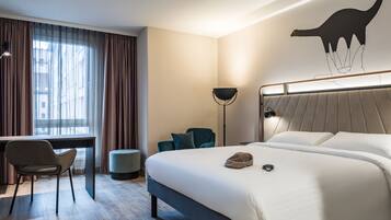 A hotel room with a bed, a desk, a chair, a lamp, and a wall art of a dinosaur.