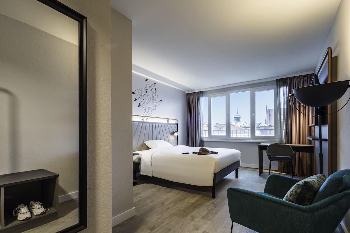 A hotel room with a bed, a desk, a chair, a large window, and a city view.