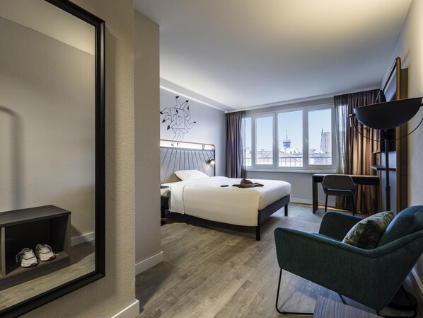 Habitación de hotel con cama, escritorio, silla, ventana grande y vistas a la ciudad.