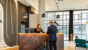 A reception desk with a modern design, a man and a woman standing, and a view of the outside through large windows. - ibis Styles Bielefeld (Bielefeld)