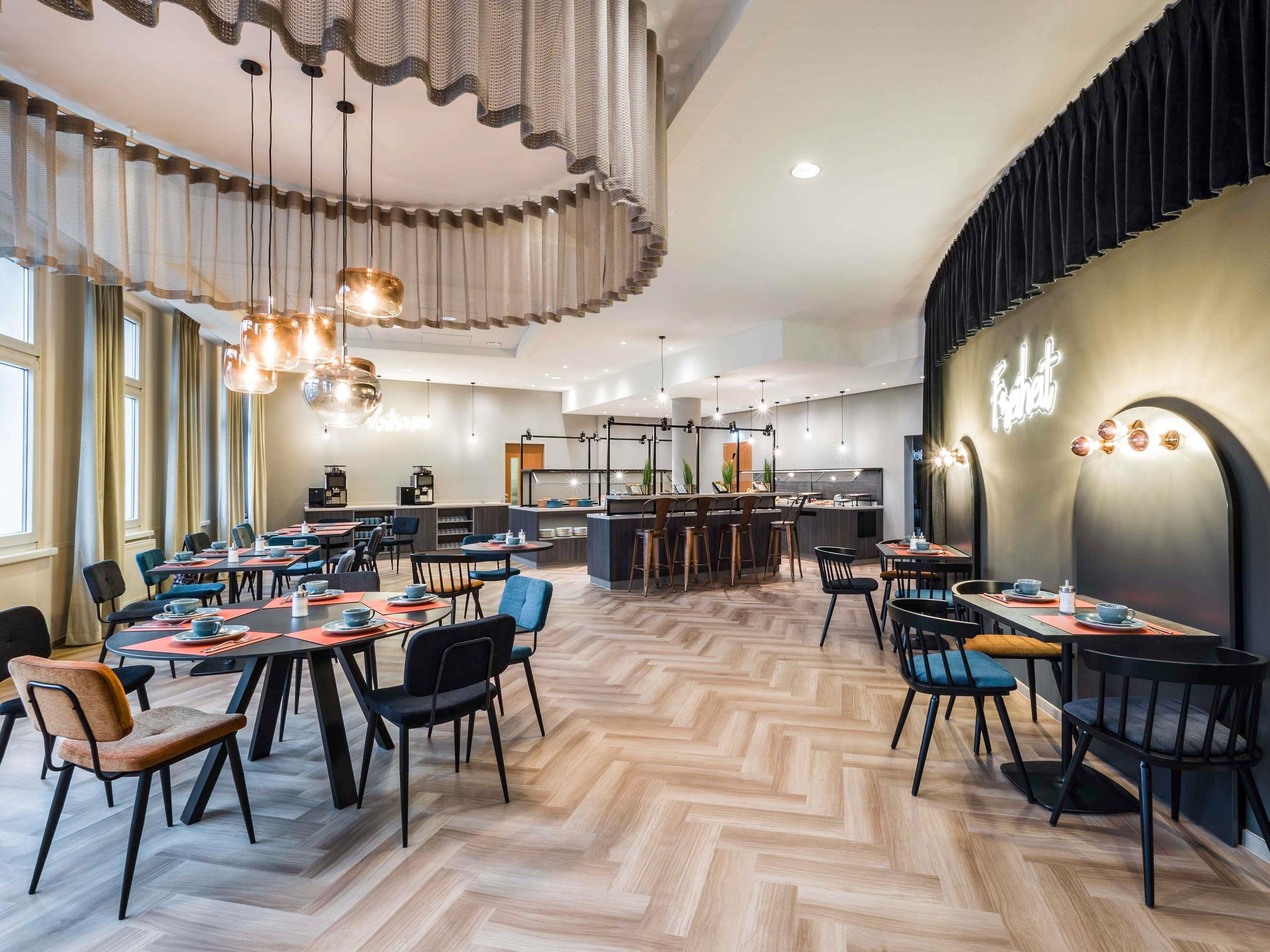 A modern restaurant with a herringbone floor, multiple dining tables, and hanging pendant lights.
