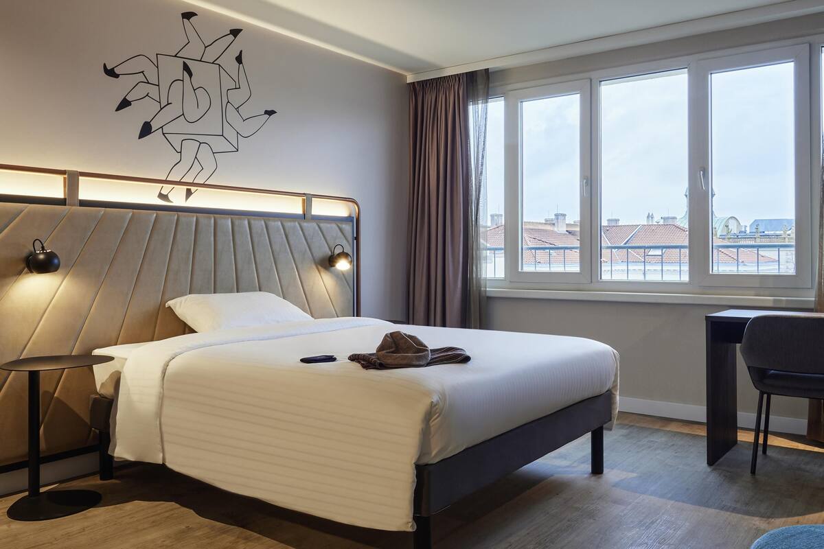 A modern hotel room with a bed, bedside lamps, a desk, and a view of the city through large windows.