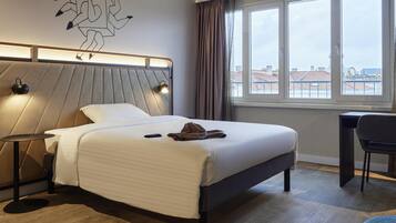 A modern hotel room with a bed, bedside lamps, a desk, and a view of the city through large windows.