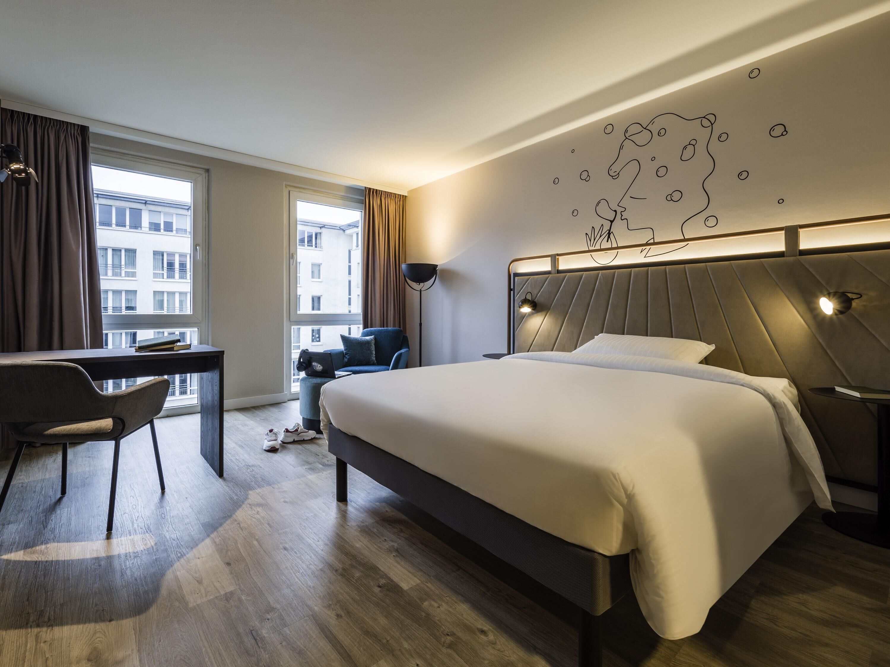 Una habitación de hotel moderna con una cama grande, un escritorio y una silla. Se ve la ciudad a través de la ventana.