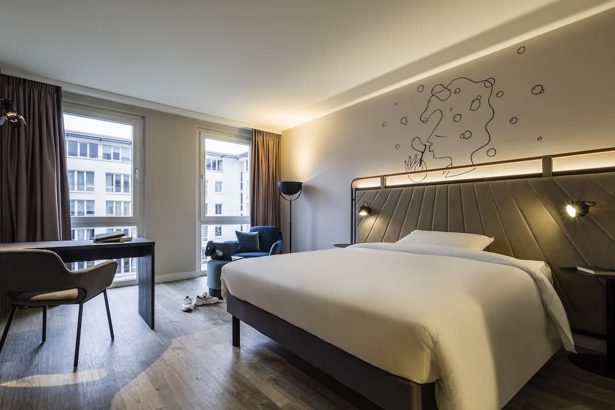 Una habitación de hotel moderna con una cama grande, un escritorio y una silla. Se ve la ciudad a través de la ventana.