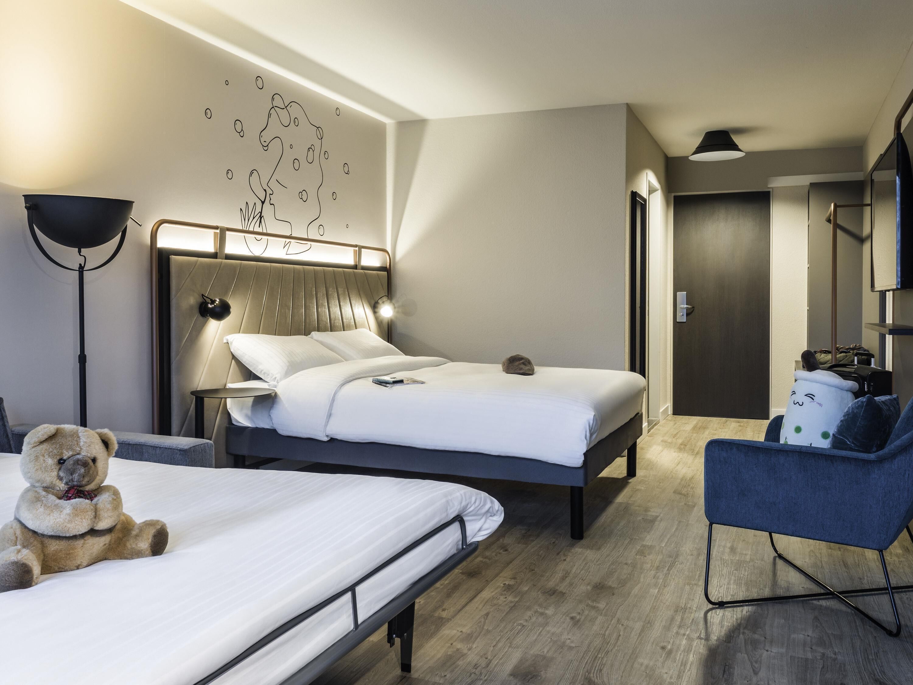 A modern hotel room with a bed, bedside table, lamp, and a teddy bear.