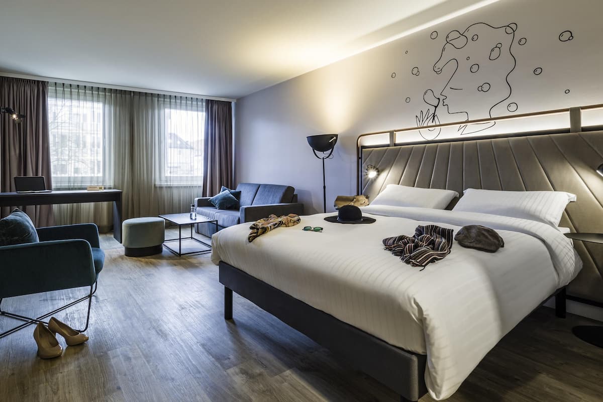 A modern hotel room with a large bed, a desk, a sofa, and a wall art piece.