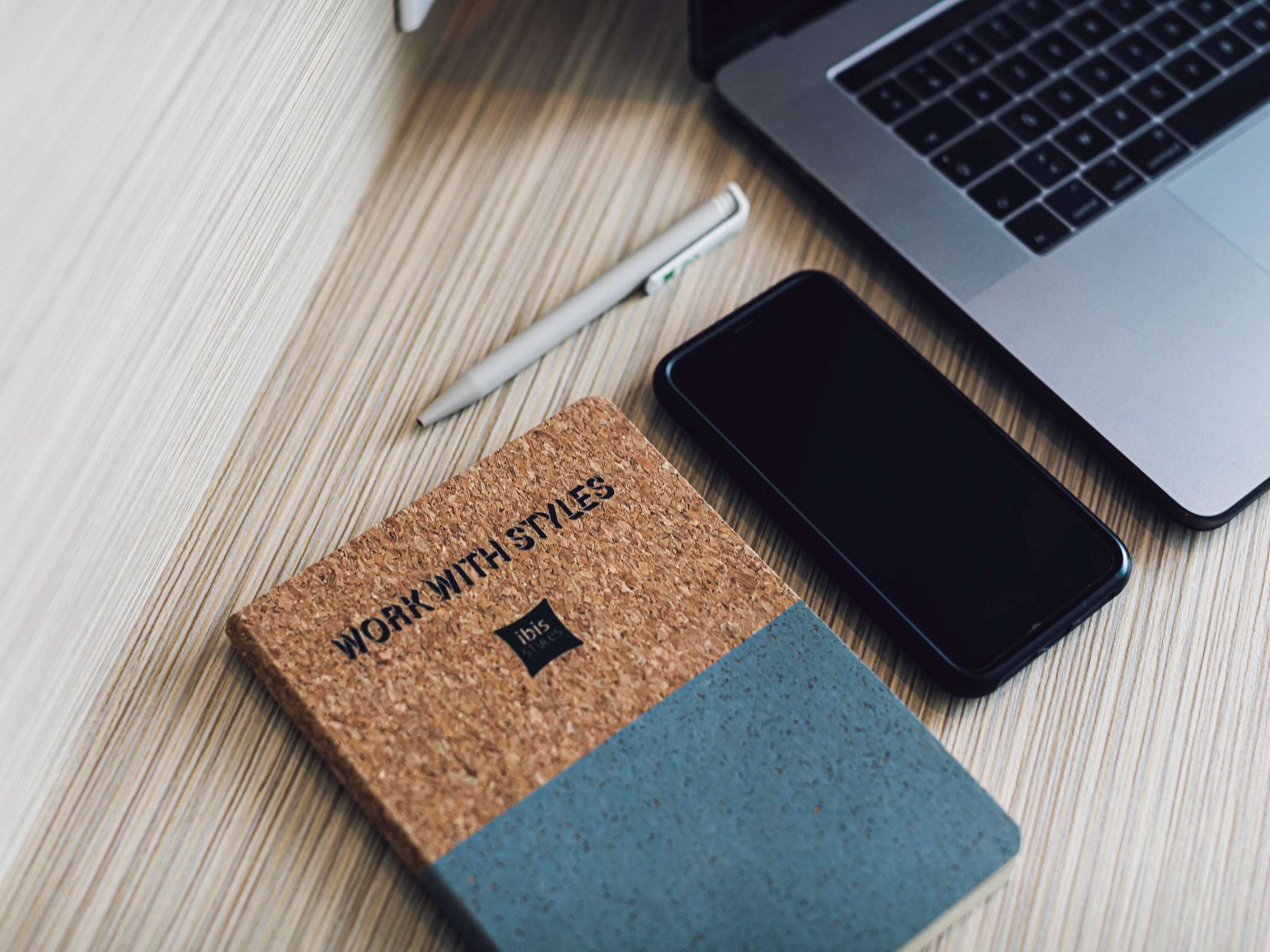 Ein Kork-Notizbuch mit der Aufschrift „WORK WITH STYLES“, ein Smartphone und ein Stift auf einer Holztischfläche.