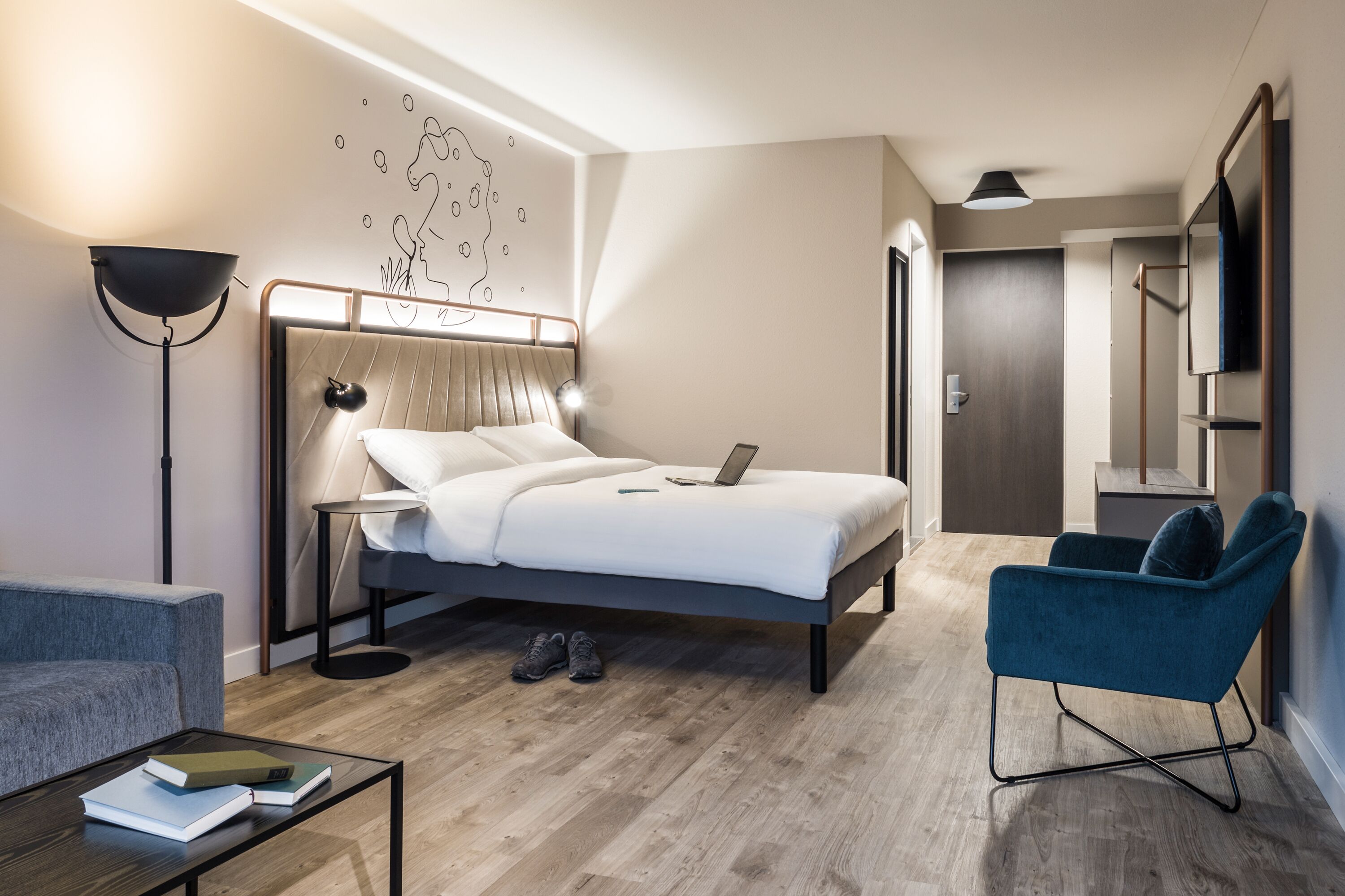 A modern hotel room with a bed, a desk, a chair, and a map on the wall.