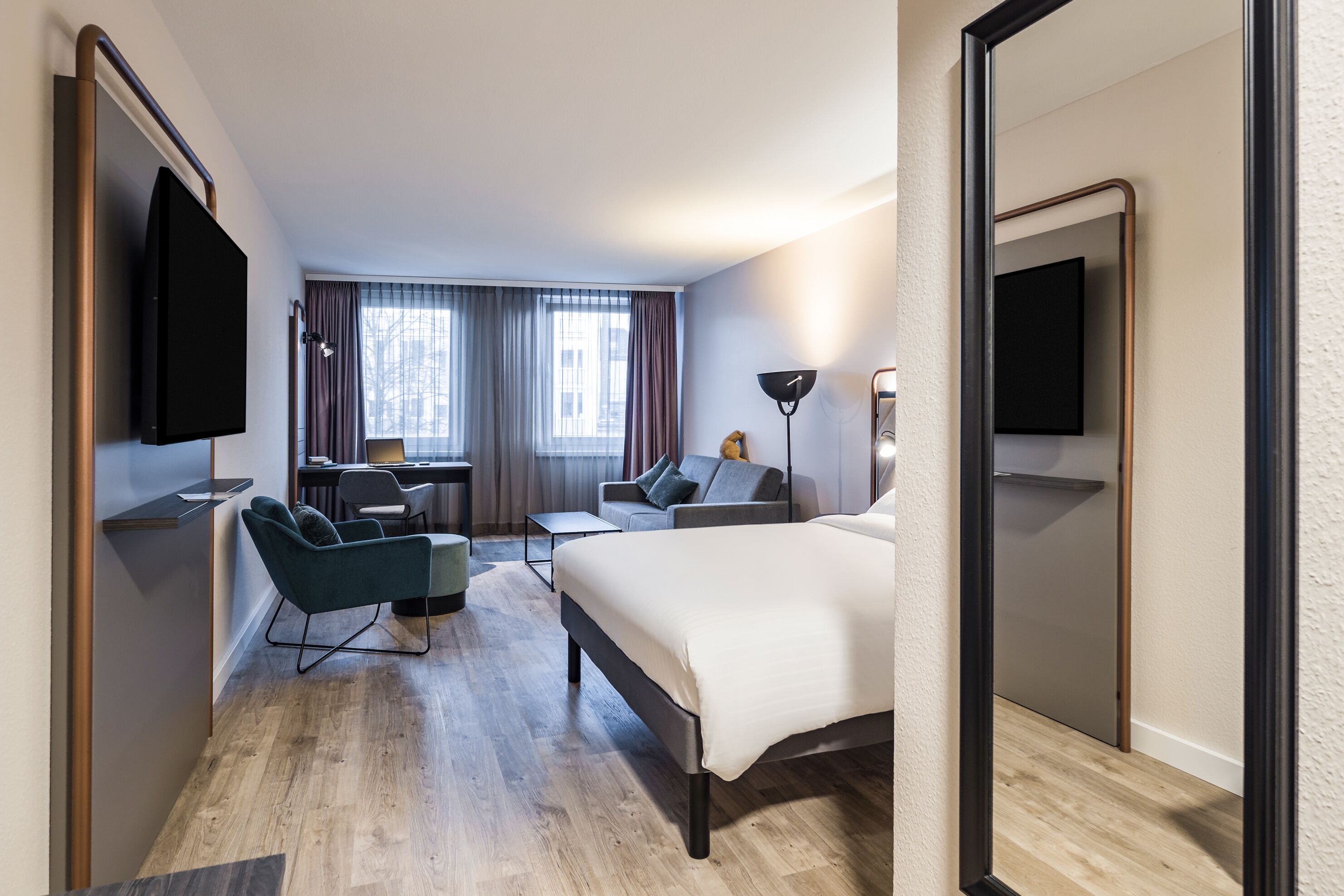 A hotel room with a bed, a desk, a chair, a sofa, a TV, and a window with curtains.