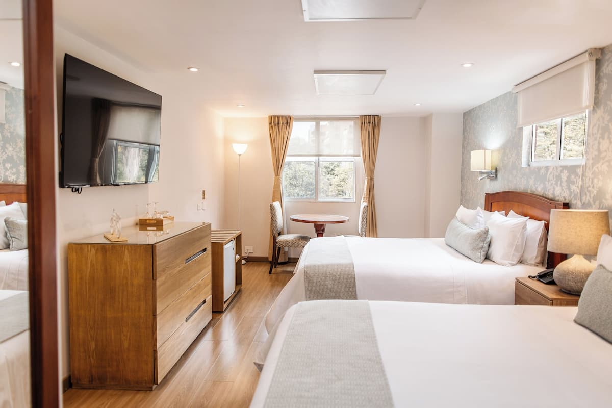 Habitación de hotel con dos camas, televisión, escritorio con silla, lámpara y ventana con cortinas.