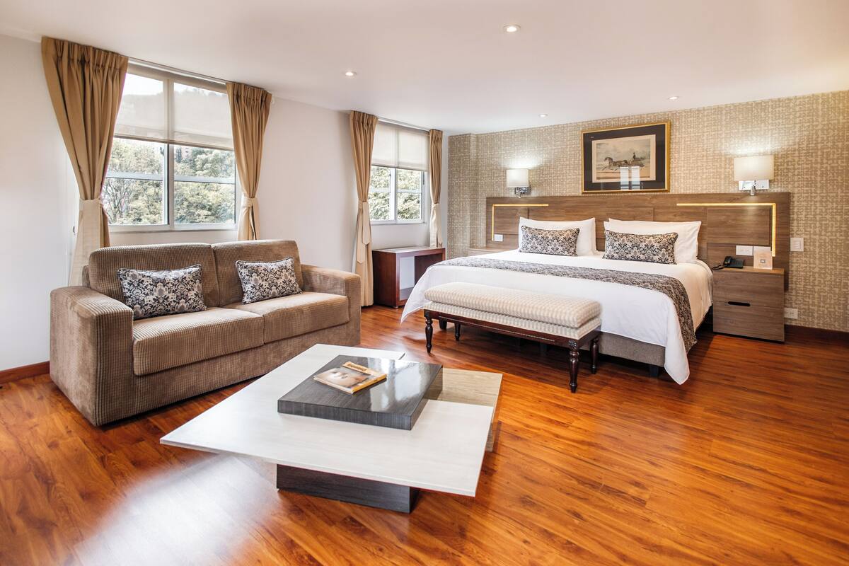 Habitación de hotel moderna con una cama grande, un sofá, una mesa de centro y vistas a árboles desde las ventanas.