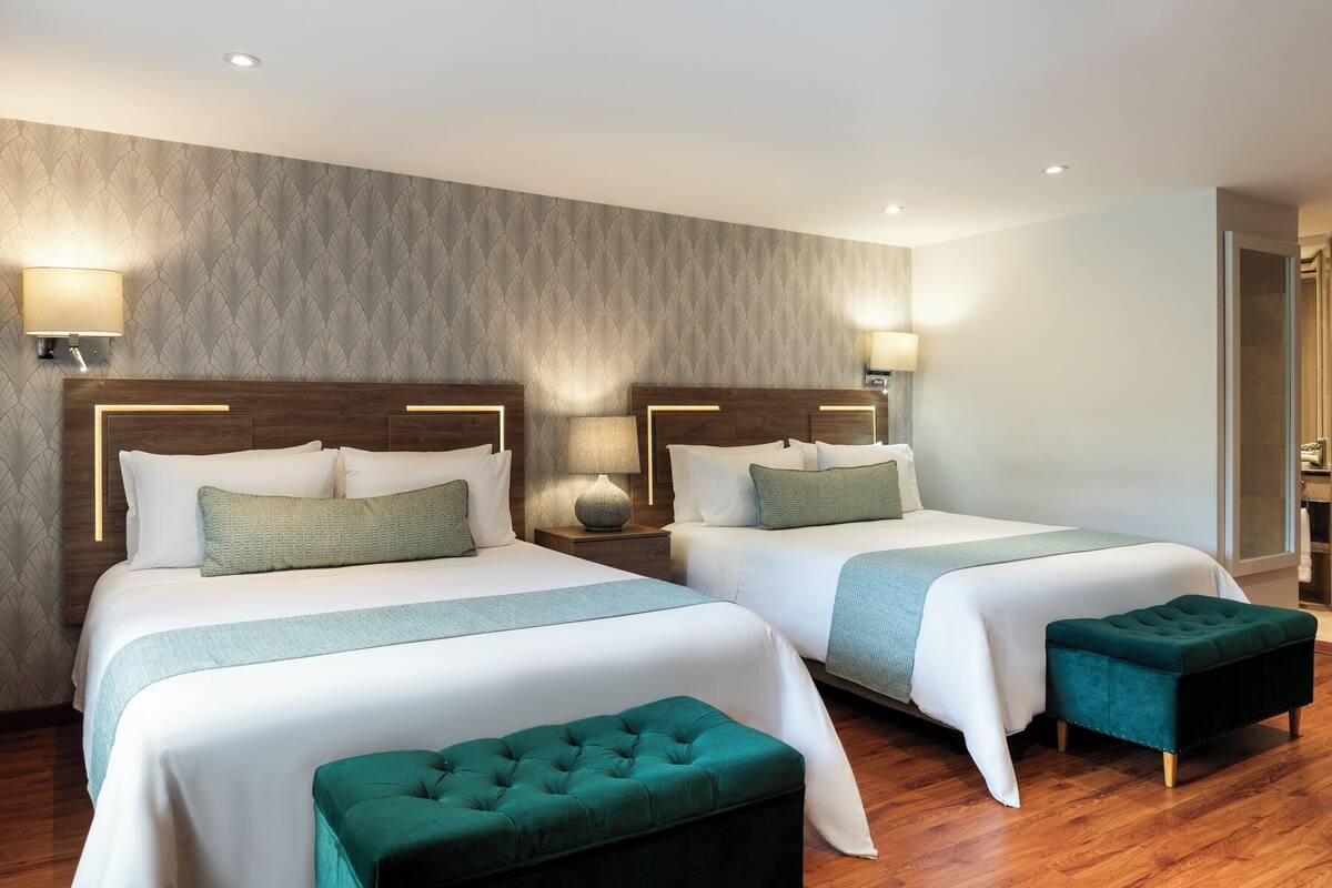 Una habitación de hotel con dos camas, cabecero de madera, lámparas de noche y un otoman verde.