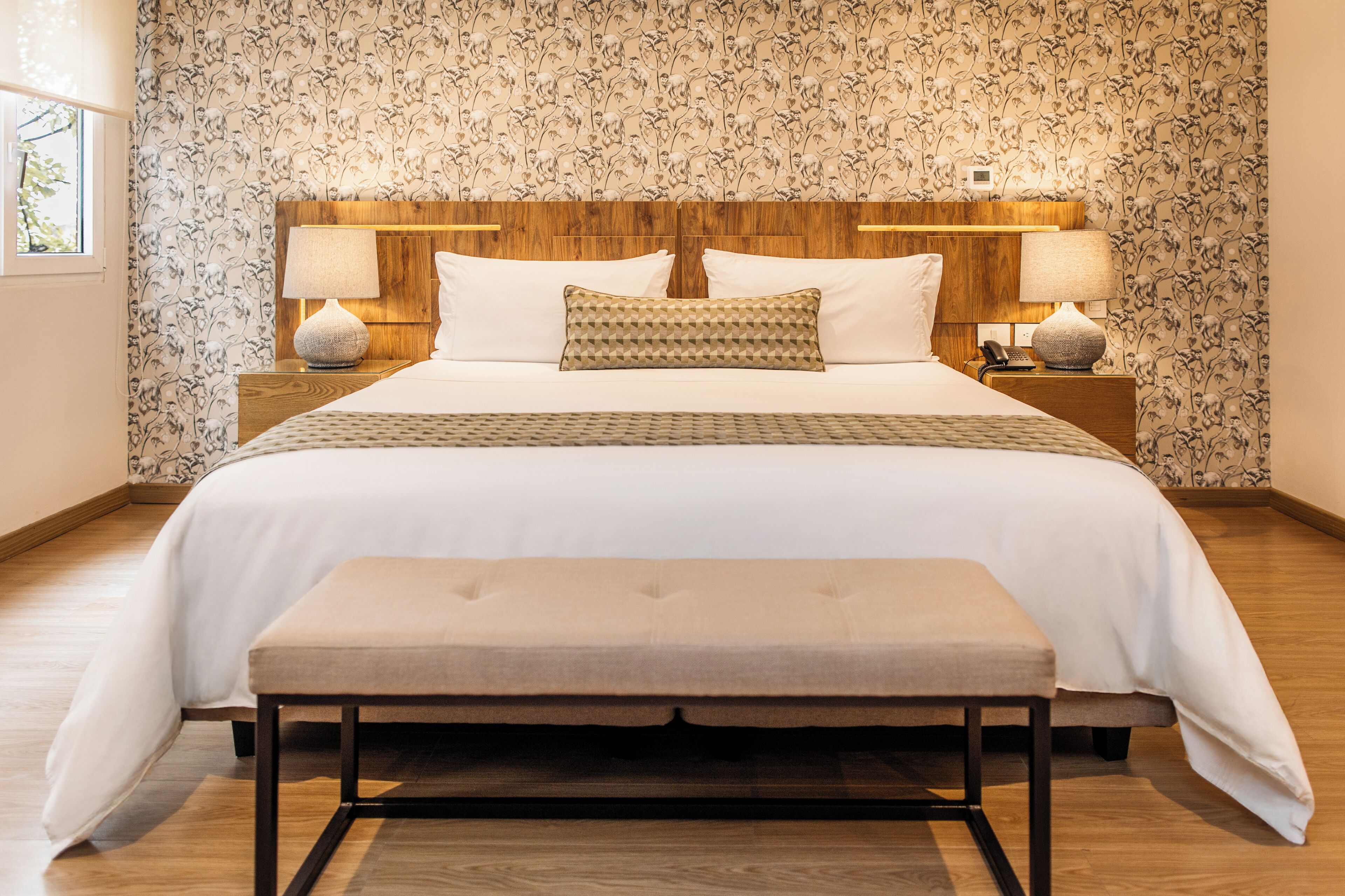 A bedroom with a wooden headboard, a large bed with white bedding, two bedside lamps, and a bench at the foot of the bed.