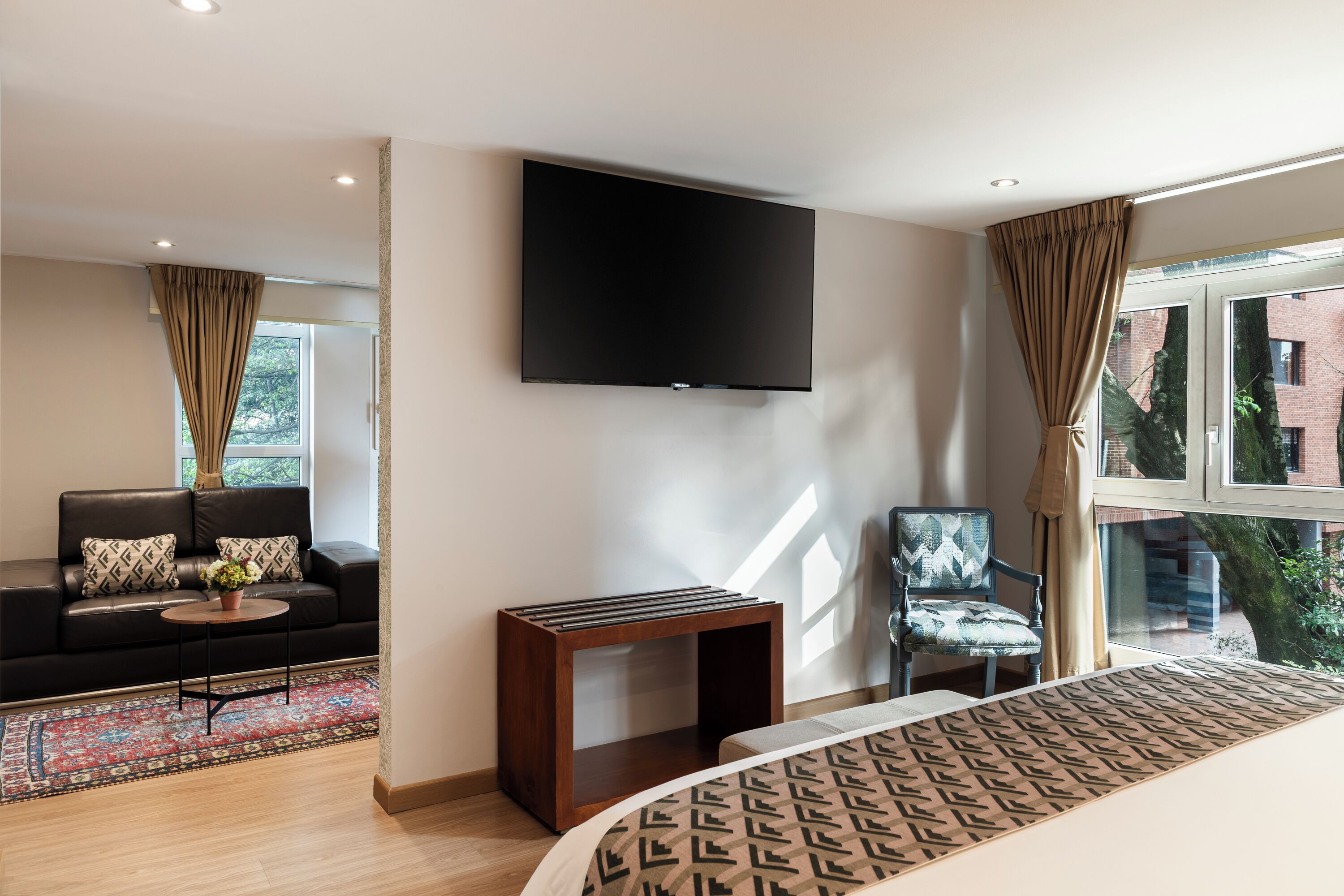 Quarto de hotel moderno com TV de tela plana, sofá, cadeira, mesa pequena e colcha estampada.