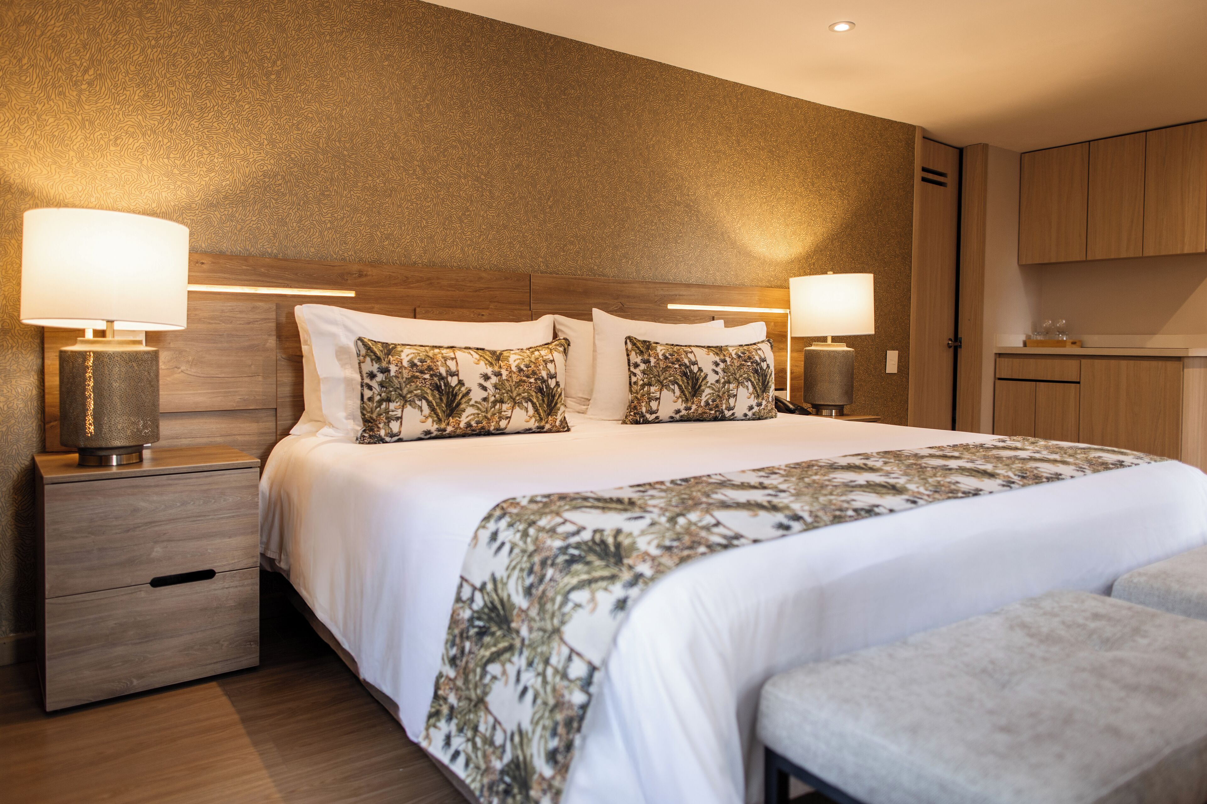 Una habitación de hotel con una cama grande, dos mesitas de noche con lámparas, un cabecero de madera y una pequeña zona de kitchenette.