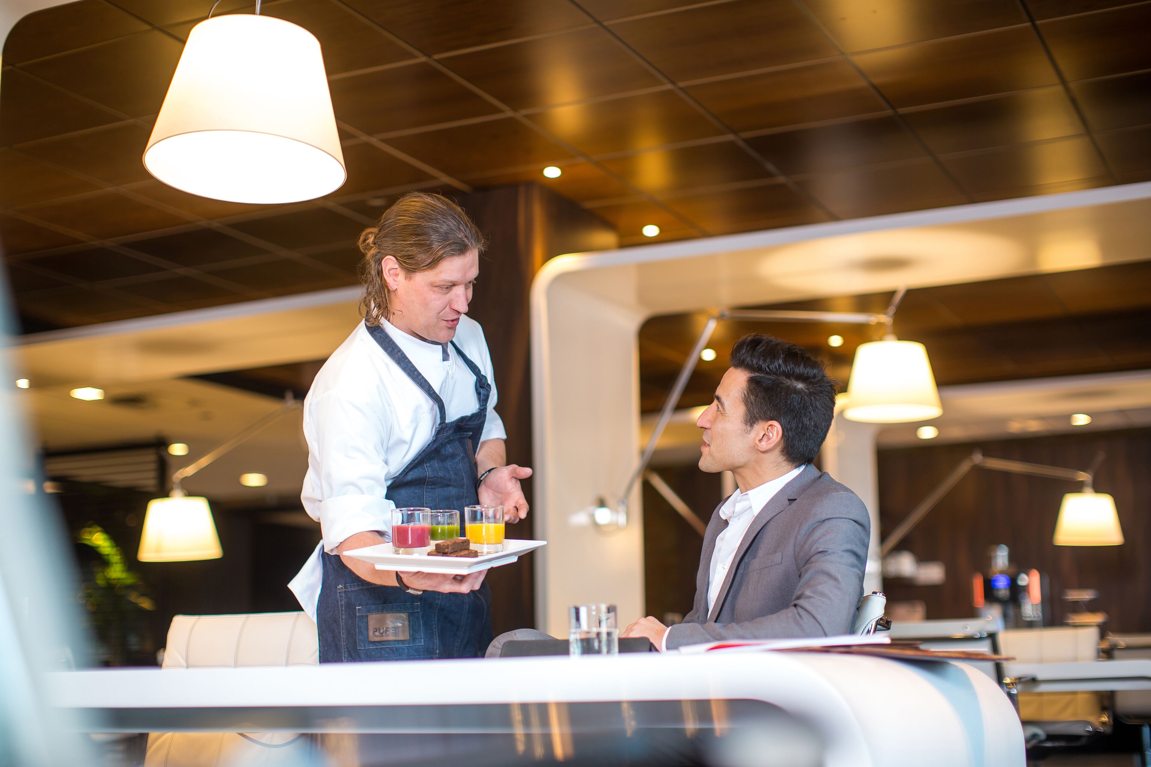 Un camarero sirve una bandeja con bebidas a un cliente sentado en un restaurante moderno con lámparas colgantes.