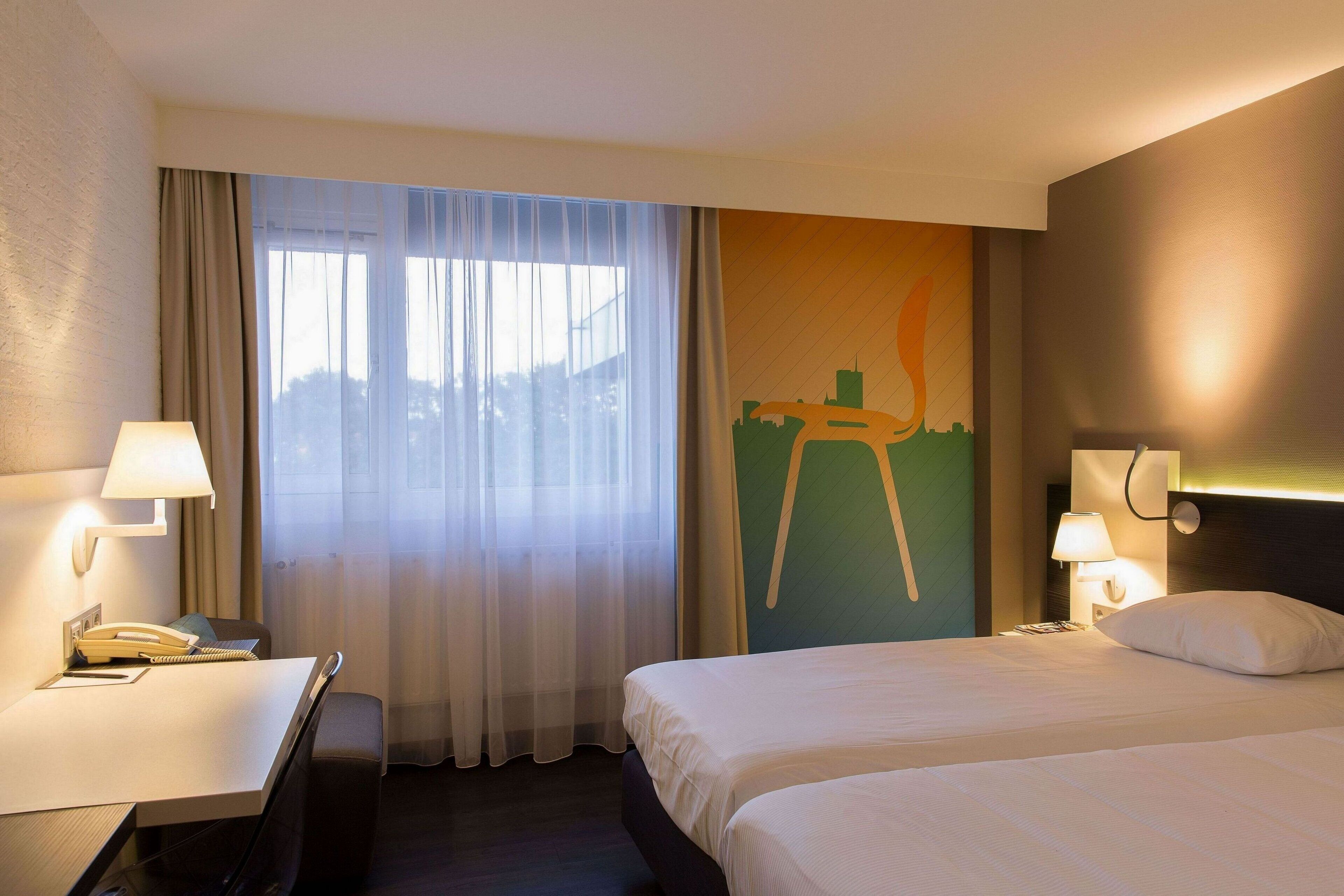 A hotel room with a bed, a desk with a telephone, a lamp, and a view of a cityscape through a window with sheer curtains.