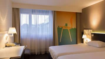 A hotel room with a bed, a desk with a telephone, a lamp, and a view of a cityscape through a window with sheer curtains.
