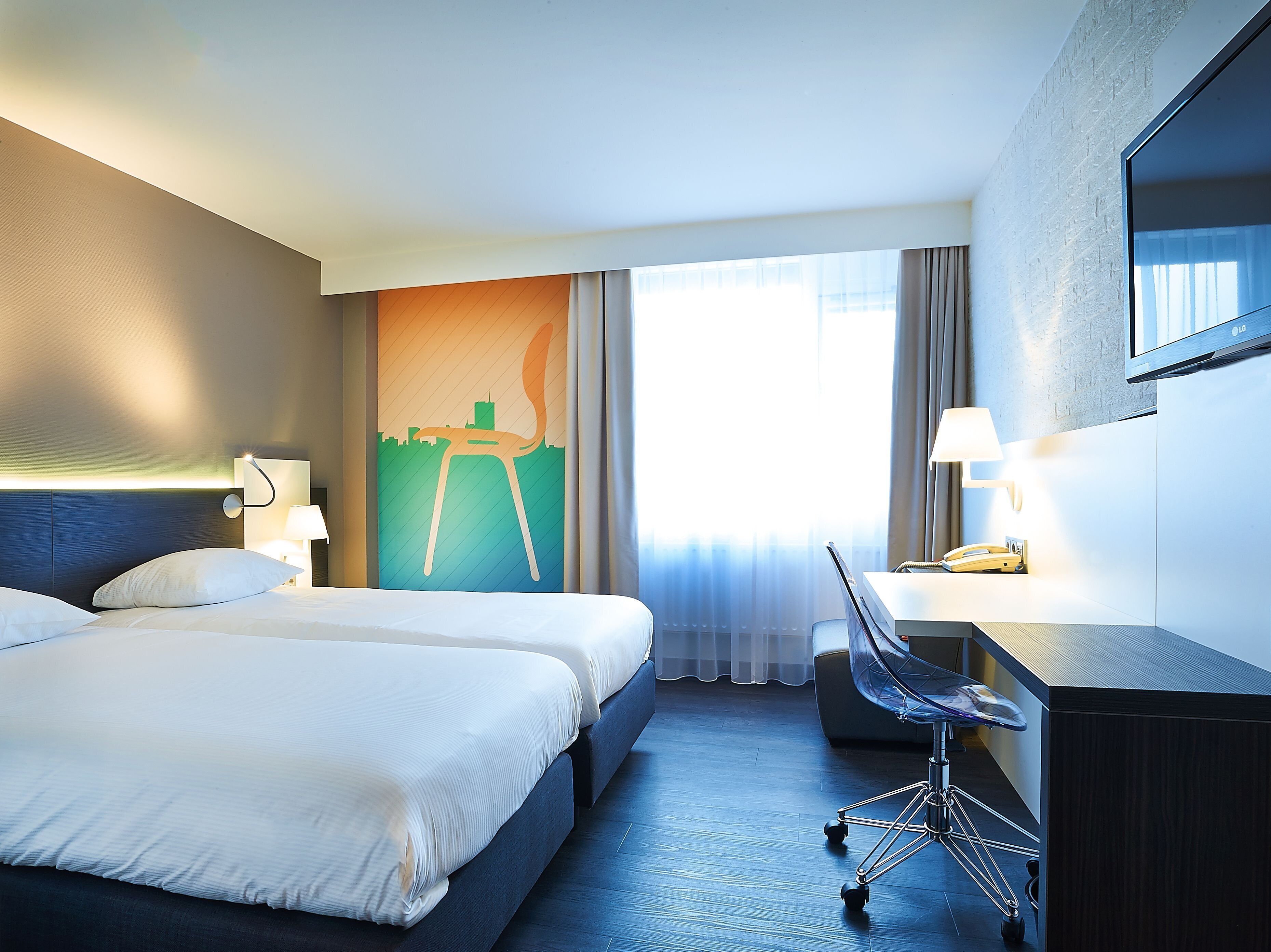 Een hotelkamer met twee bedden, een bureau, een stoel, een stadsgezicht kunstwerk en een groot raam met gordijnen.