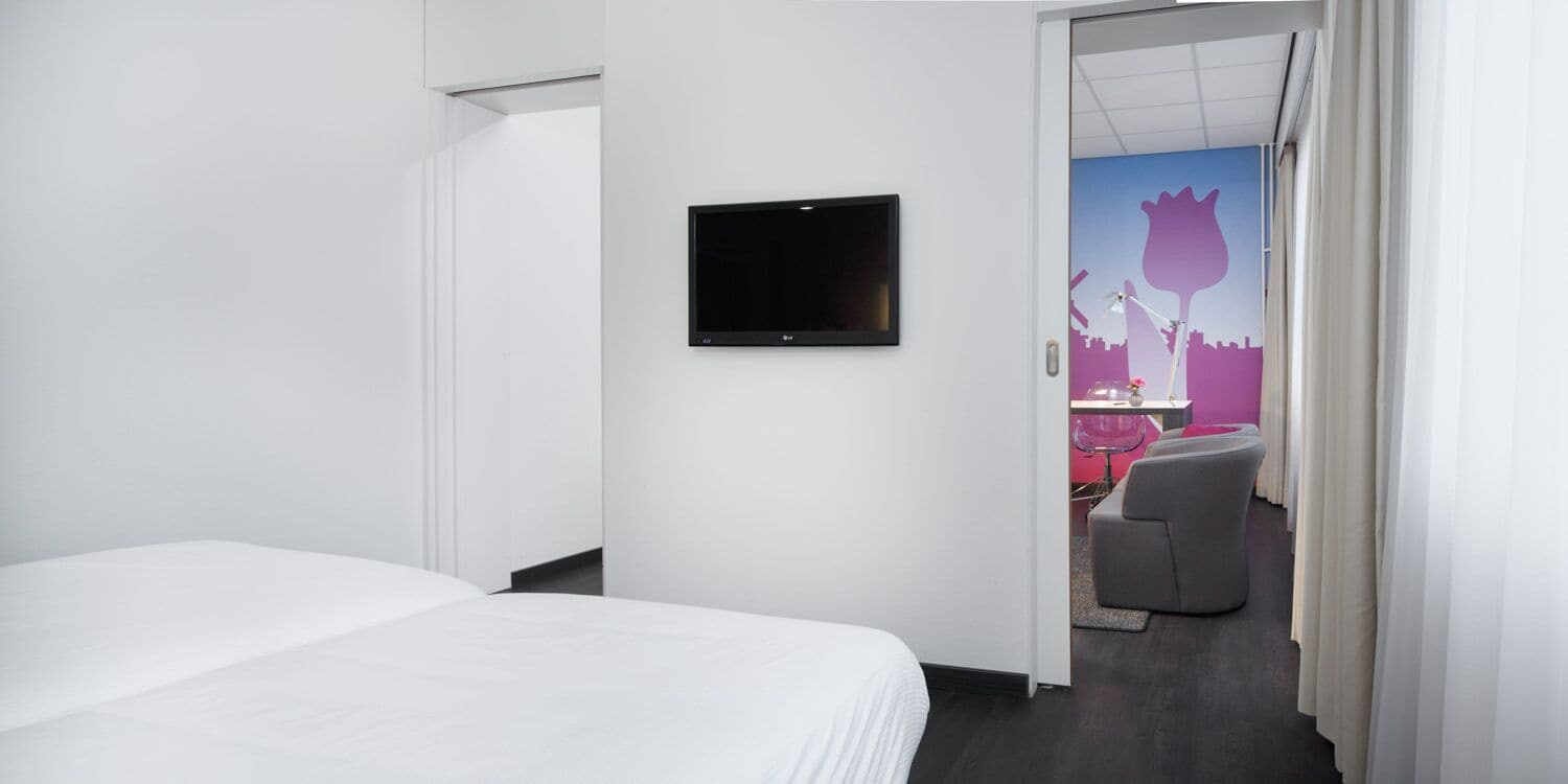 A hotel room with a bed, a TV mounted on the wall, and a view of a salon with a pink chair.