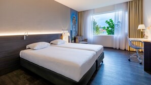 A hotel room with two beds, a desk, a chair, and a large window with a view of greenery. - Postillion Hotel Dordrecht (Dordrecht)