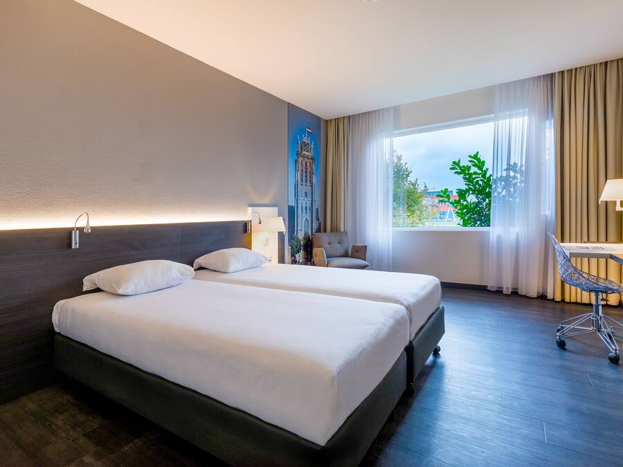 A hotel room with two beds, a desk, a chair, and a large window with a view of greenery.