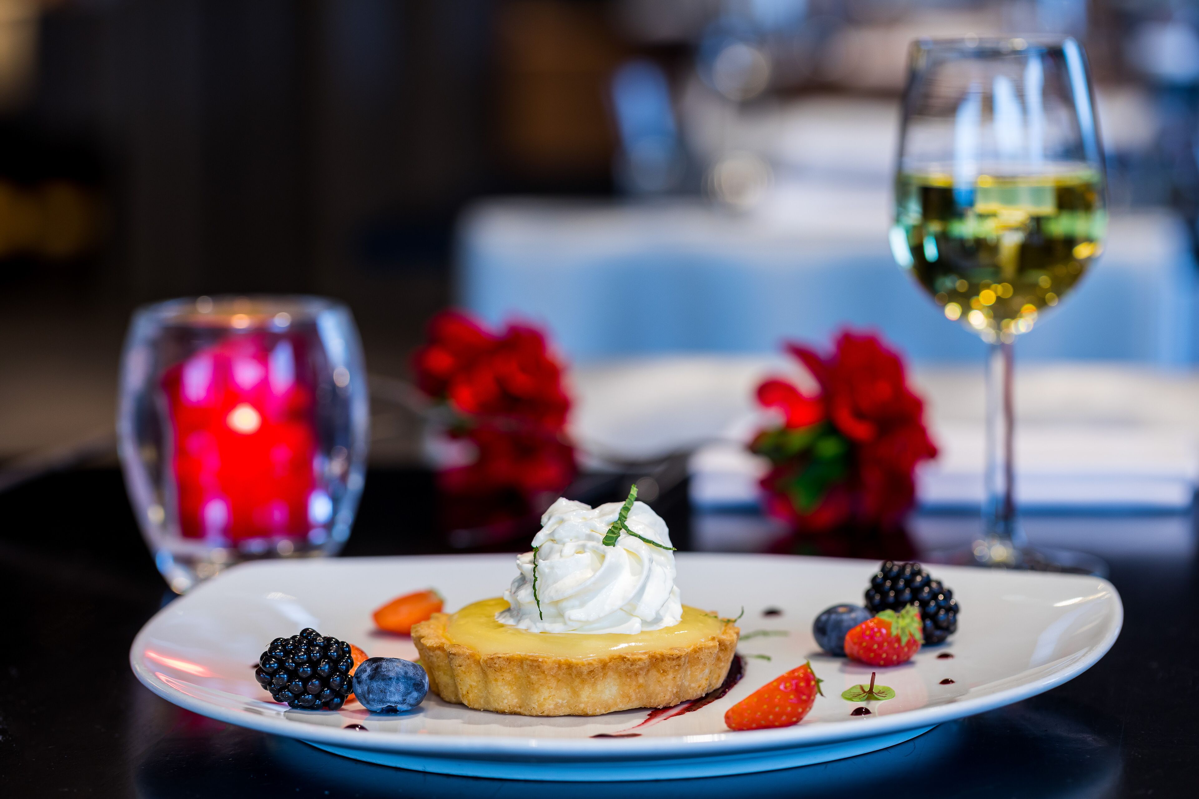 Um prato de sobremesa com torta, chantilly, frutas vermelhas e um copo de vinho branco.