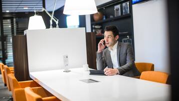 Un bureau moderne avec un bureau blanc, un ordinateur portable et une tablette. On y trouve des chaises orange et une lampe blanche.