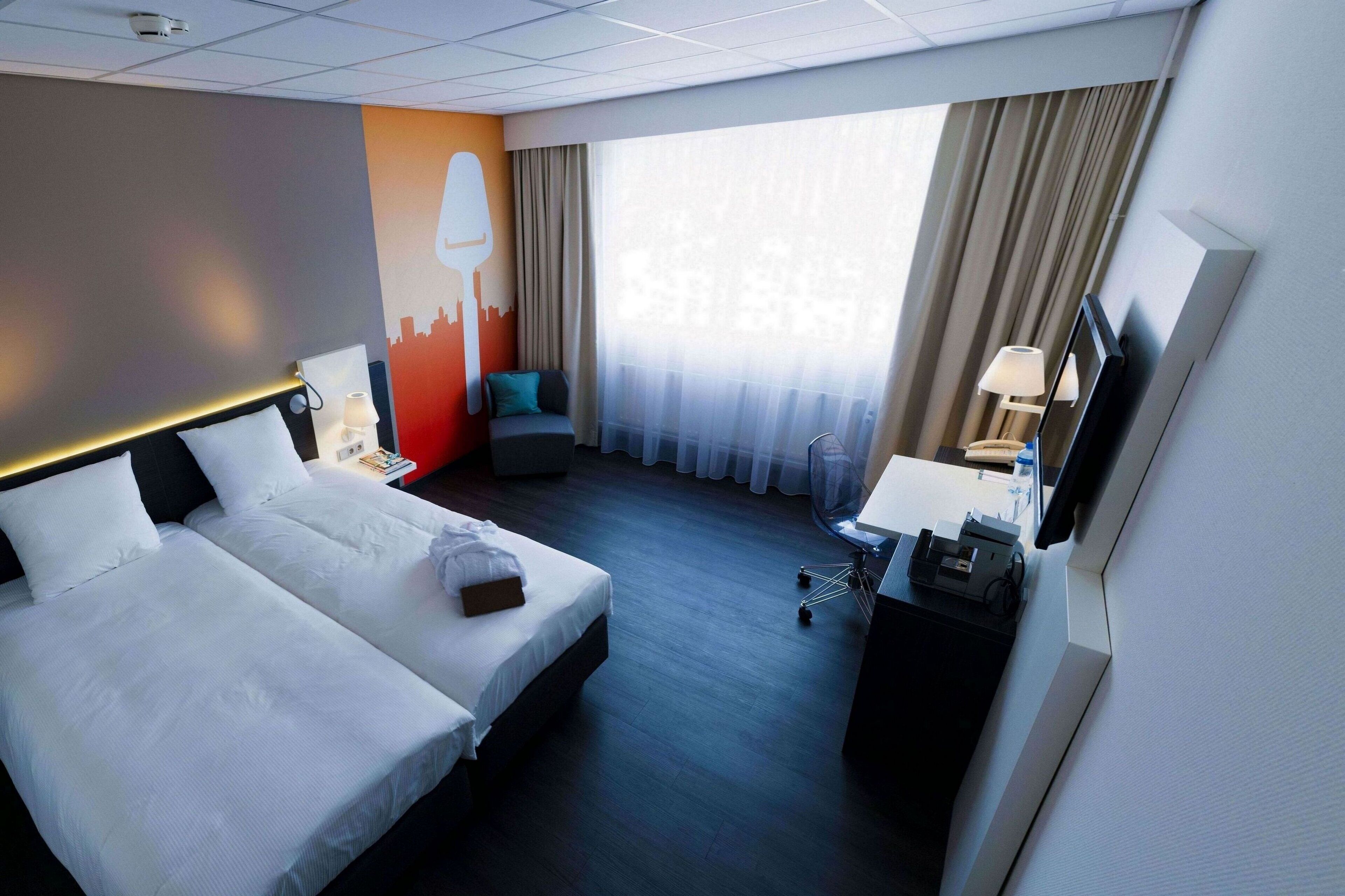 Habitación de hotel con dos camas, un mural de paisaje urbano, un ventanal con cortinas, un escritorio con lámpara y un televisor.