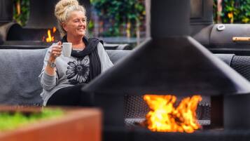 A person sitting on a grey sofa with a white mug, in front of a wood-burning stove with visible flames.