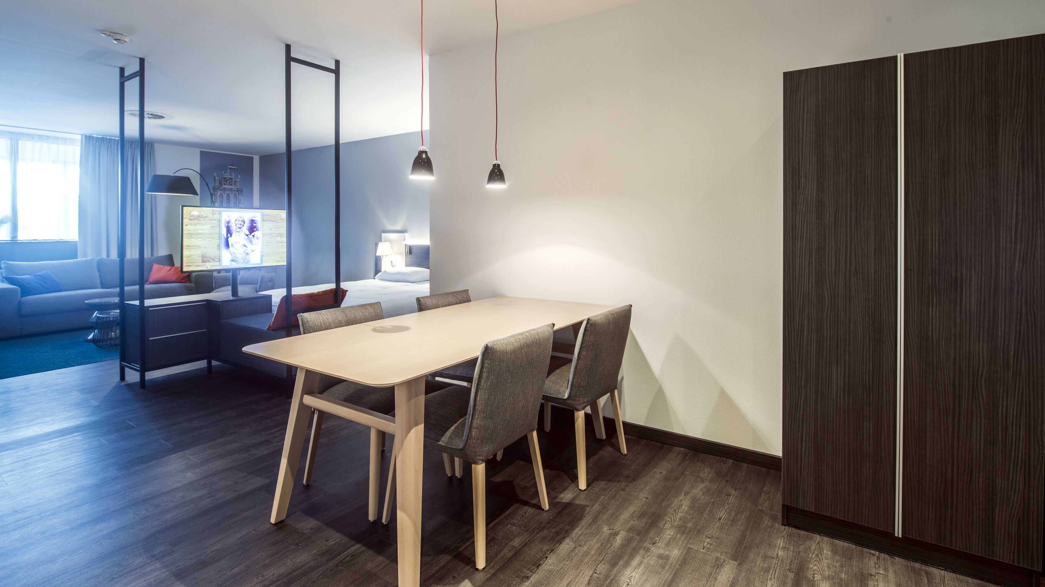 A modern hotel room with a dining area, a sofa, a TV, and a wardrobe.