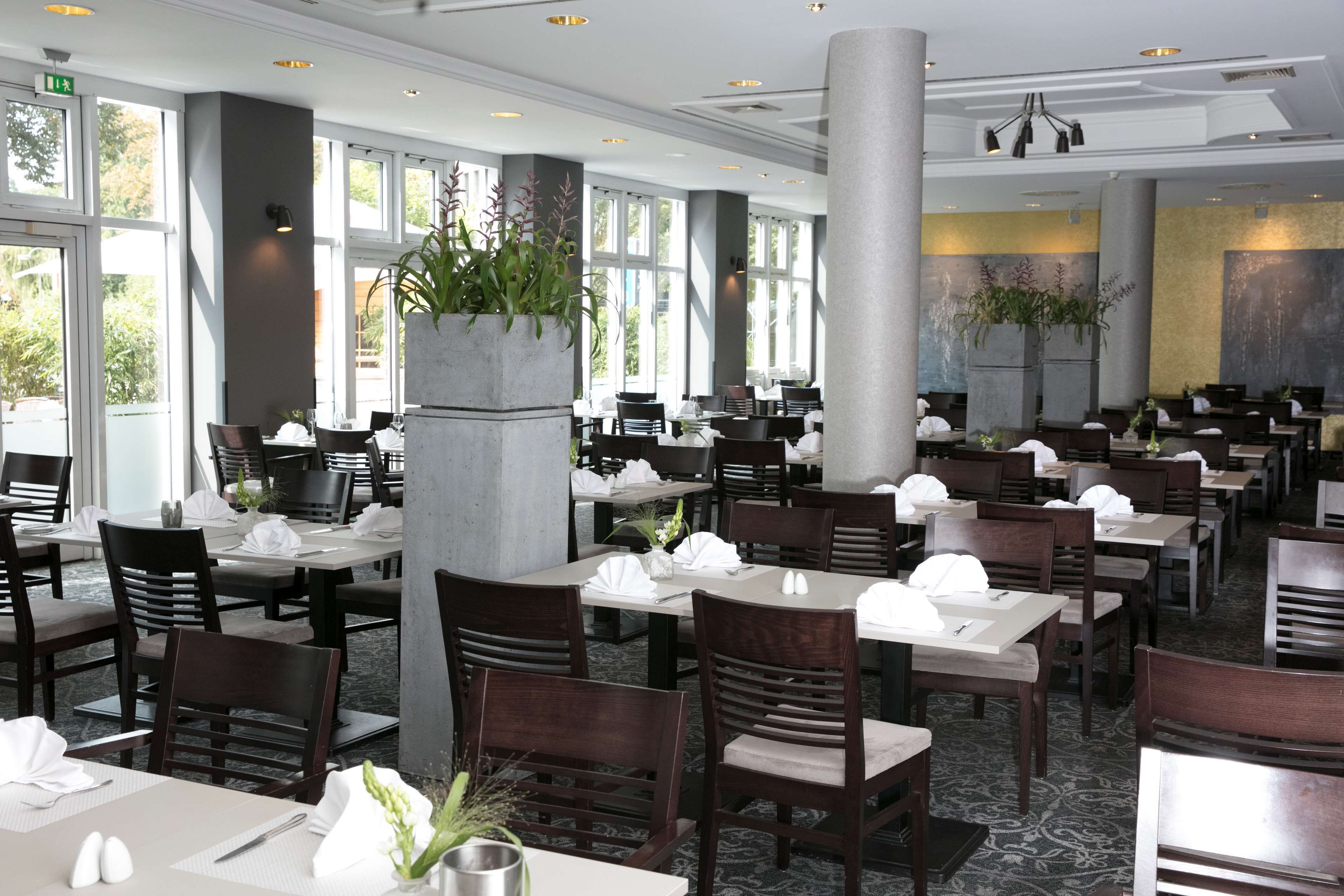 Un restaurante amplio con techos altos, ventanas grandes y decoración moderna. Las mesas están dispuestas ordenadamente con manteles blancos y sillas.