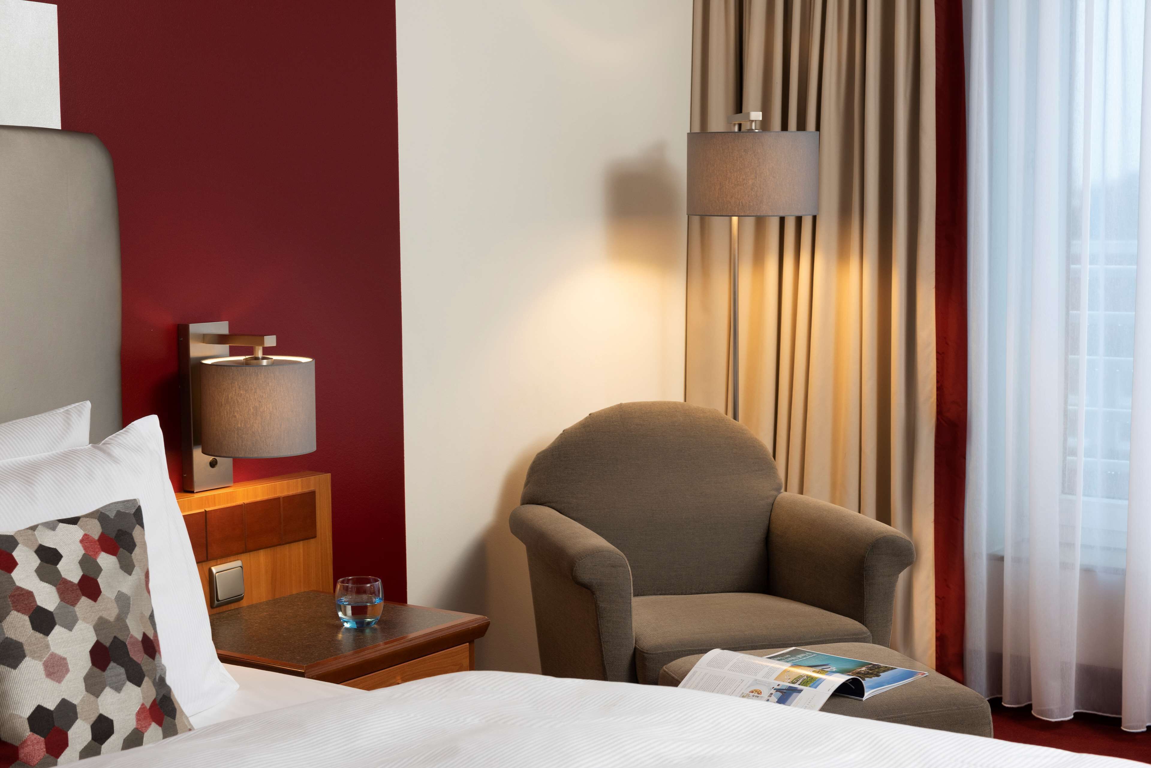 Habitación de hotel con cama, una silla, una mesita de noche con lámpara y ventana con cortinas.
