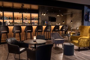 Uma área de bar com vários bancos e balcão, uma área de estar com mesas redondas e cadeiras, e uma parede com uma tela de televisão.