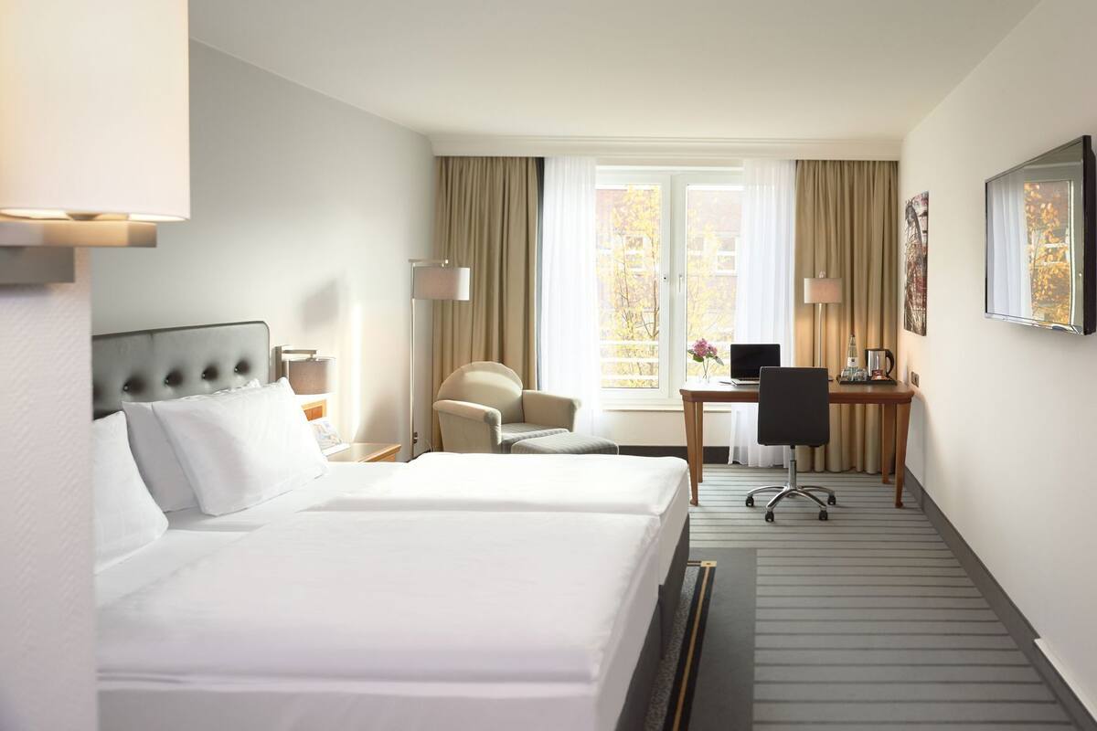 A hotel room with two beds, a desk, a chair, a lamp, and a window with curtains.