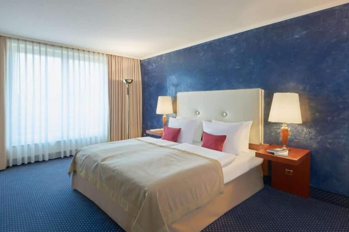 A hotel room with a large bed, two bedside tables with lamps, a window with curtains, and a blue textured wall.