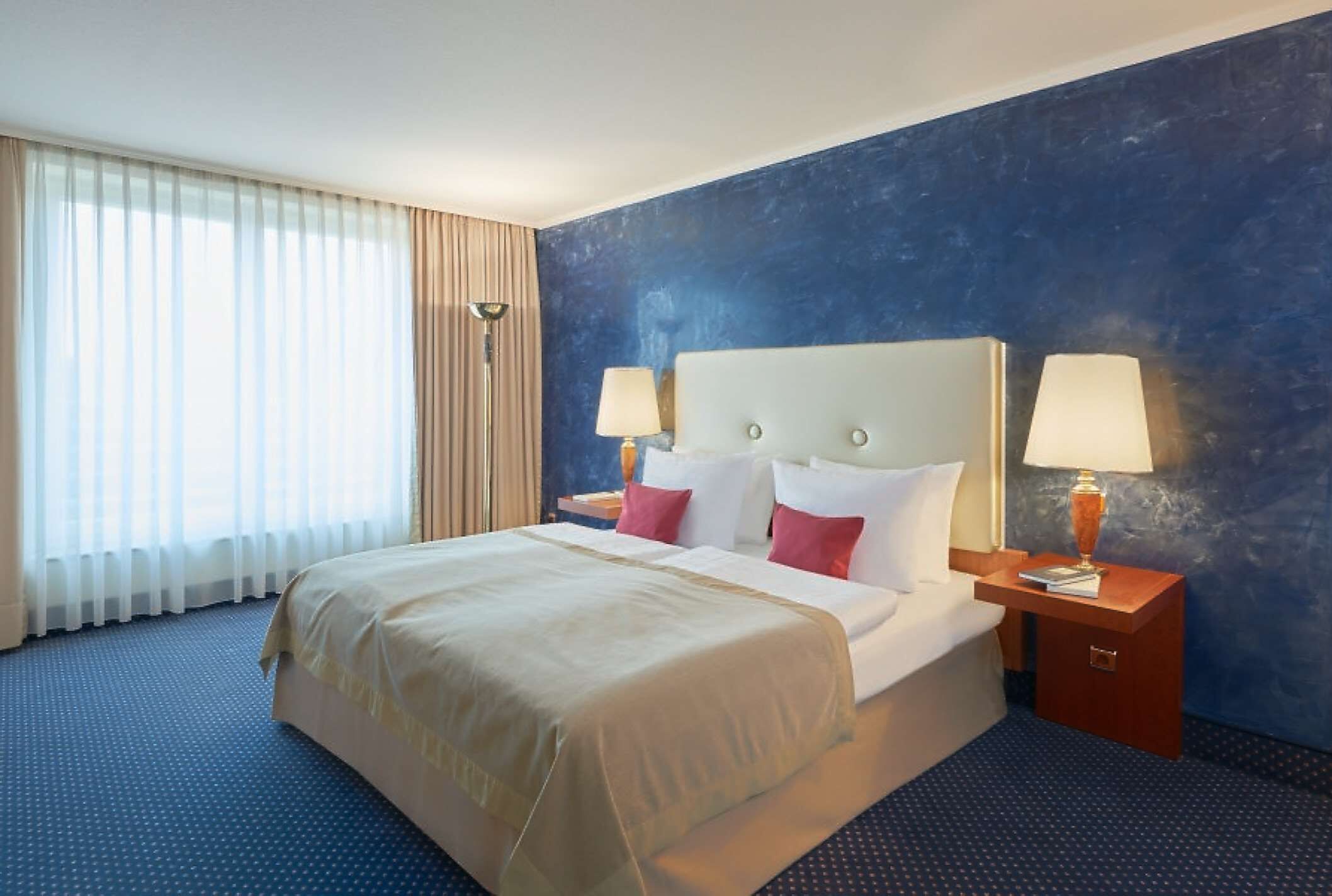 Ein Hotelzimmer mit einem großen Bett, zwei Nachttischen mit Lampen, einem Fenster mit Vorhängen und einer Wand in Blautönen.