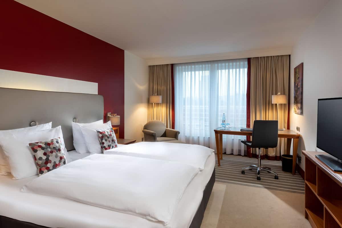 A hotel room with two beds, a desk, a chair, a TV, and a window with curtains.