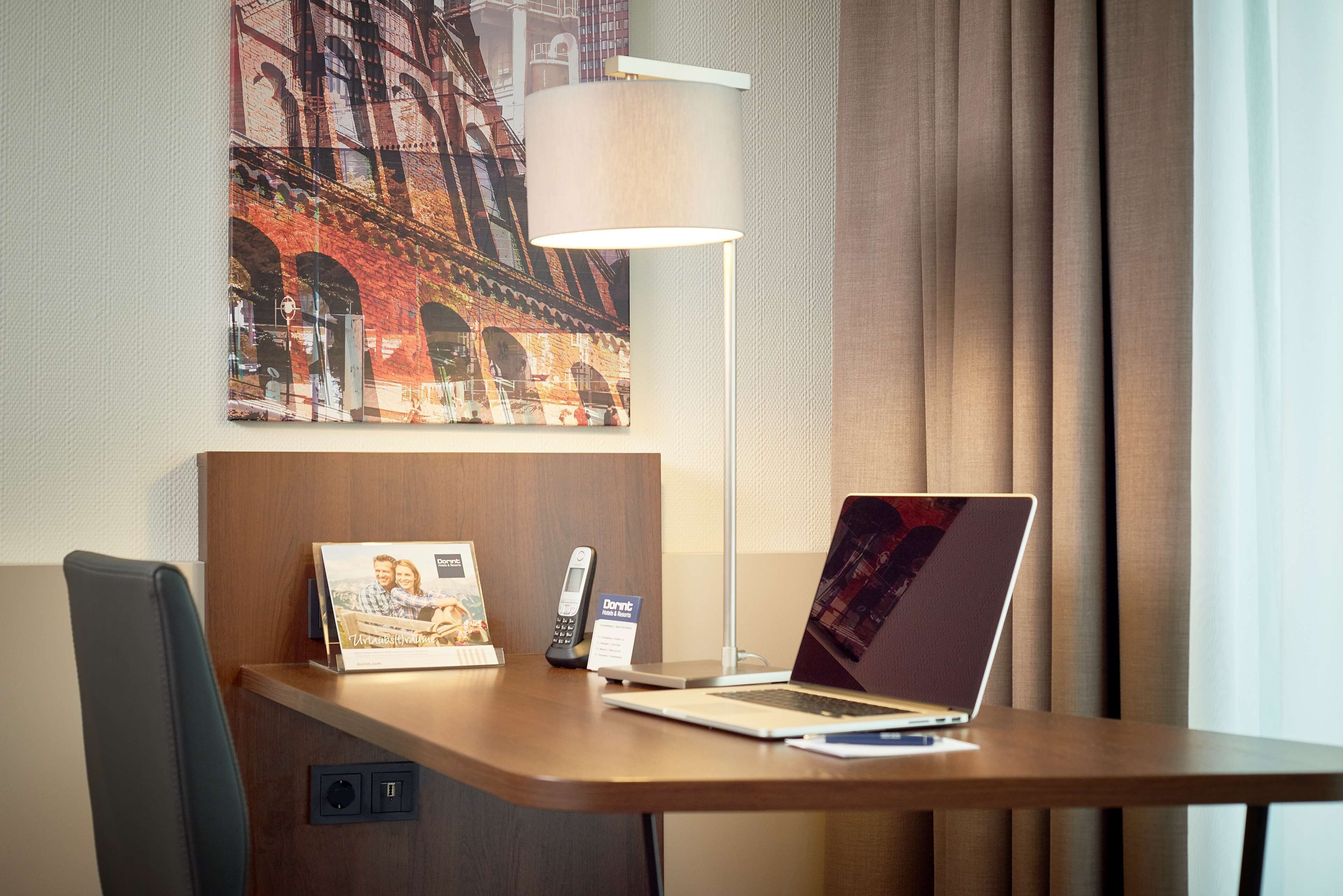 A hotel room desk with a laptop, a phone, a lamp, and a framed photo.