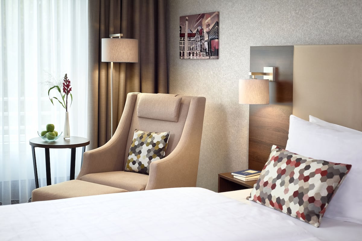 A hotel room with a bed, a sofa, a small table with a vase and fruit, and a wall with a painting and two wall-mounted lamps.