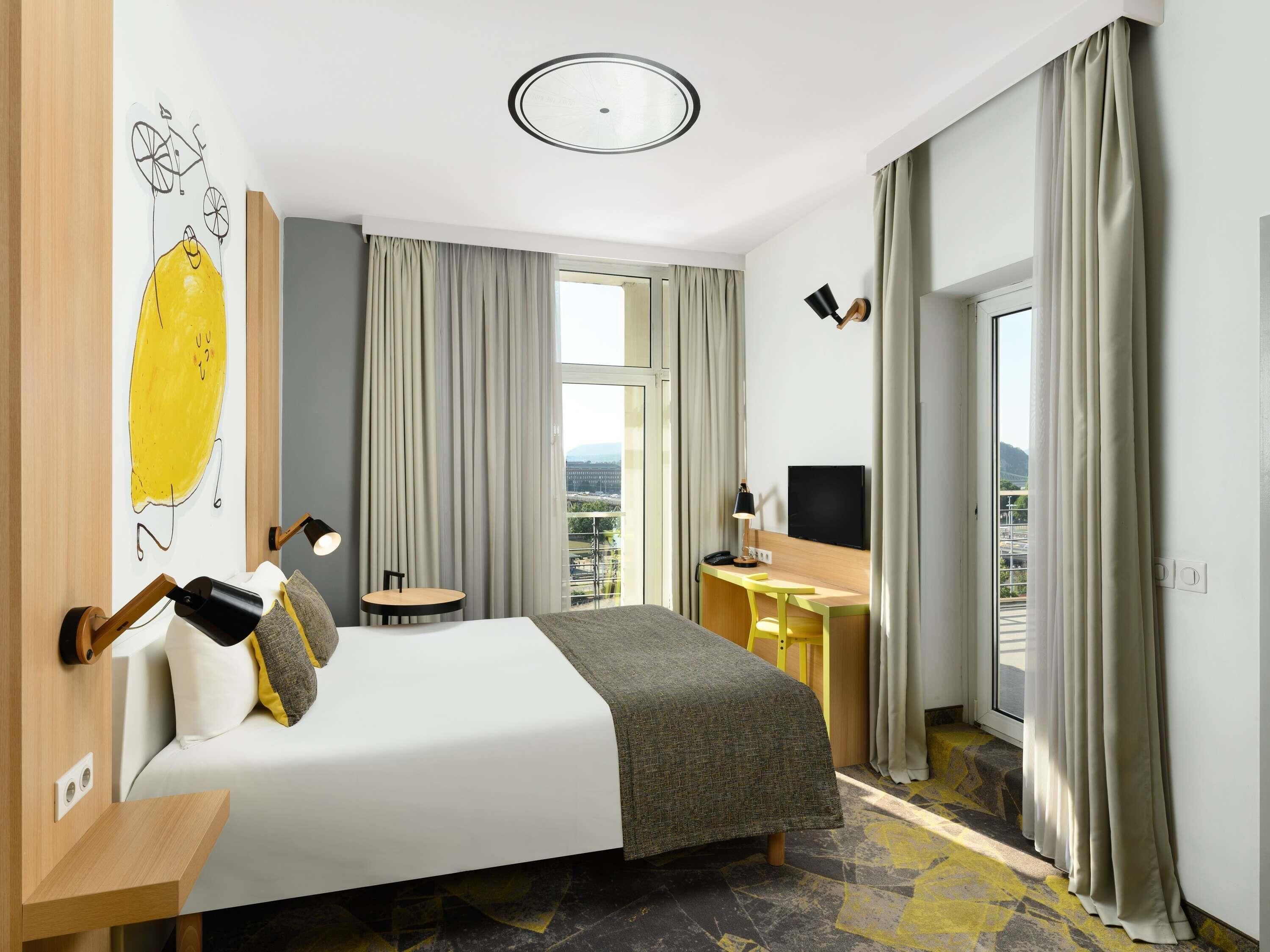 A hotel room with a bed, desk, and a large window with curtains.