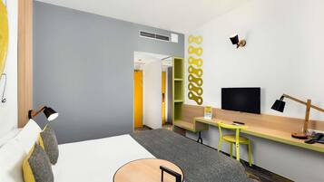 A modern hotel room with a bed, a desk, a chair, a television, and a wall-mounted shelf.