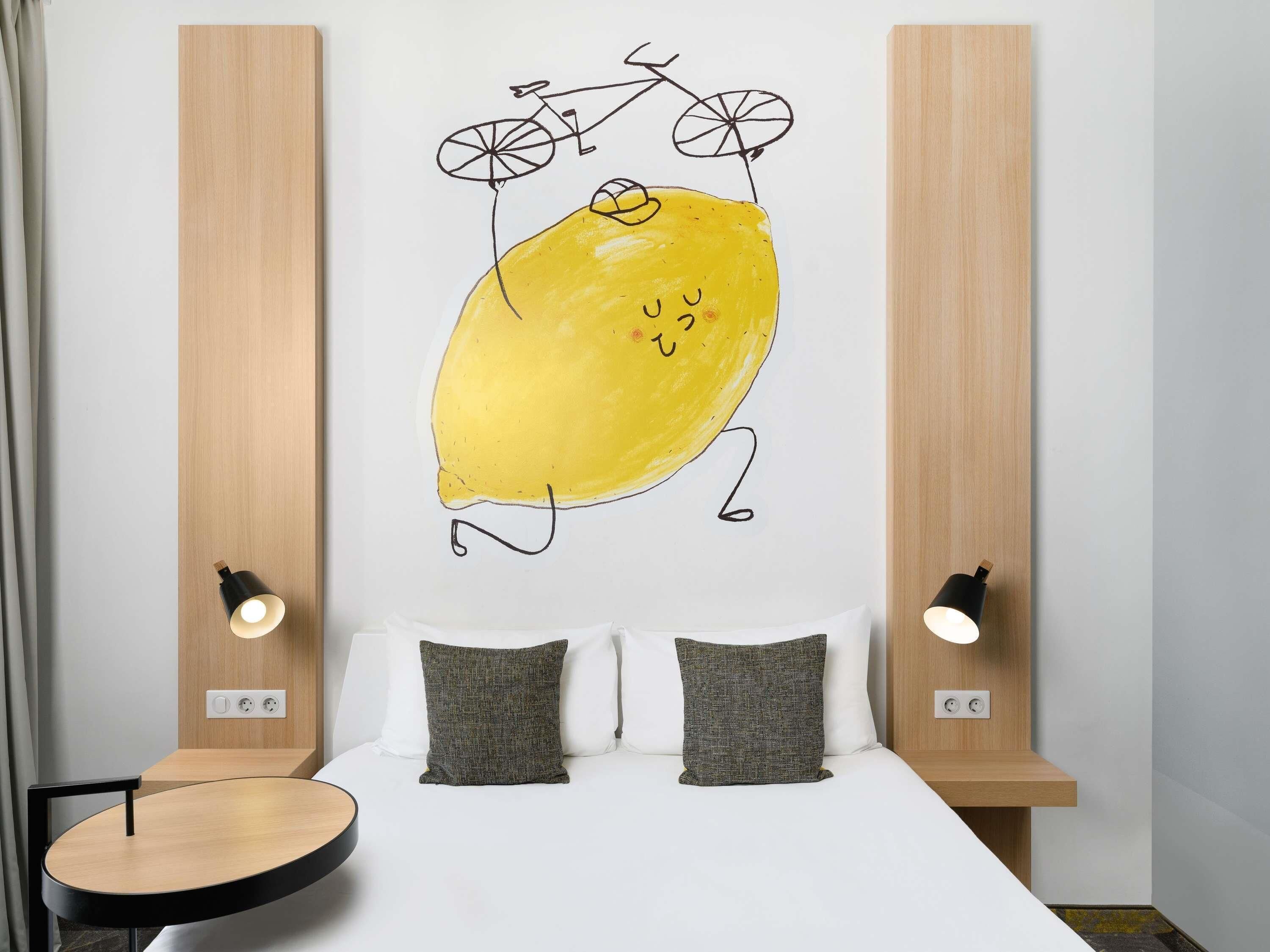 Un dormitorio con cama, dos almohadas, una lámpara de pared y una obra de arte en la pared de un limón y una bicicleta.