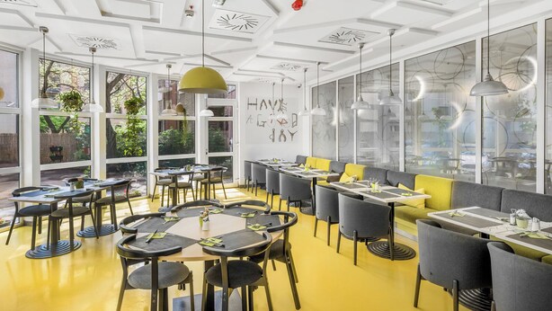 Un restaurante moderno con mesas redondas, sillas amarillas y grises, y amplias ventanas con vistas al exterior.