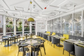 A modern restaurant with round tables, yellow and grey chairs, and large windows offering views of the outdoors. - ibis Styles Budapest City (Budapest)