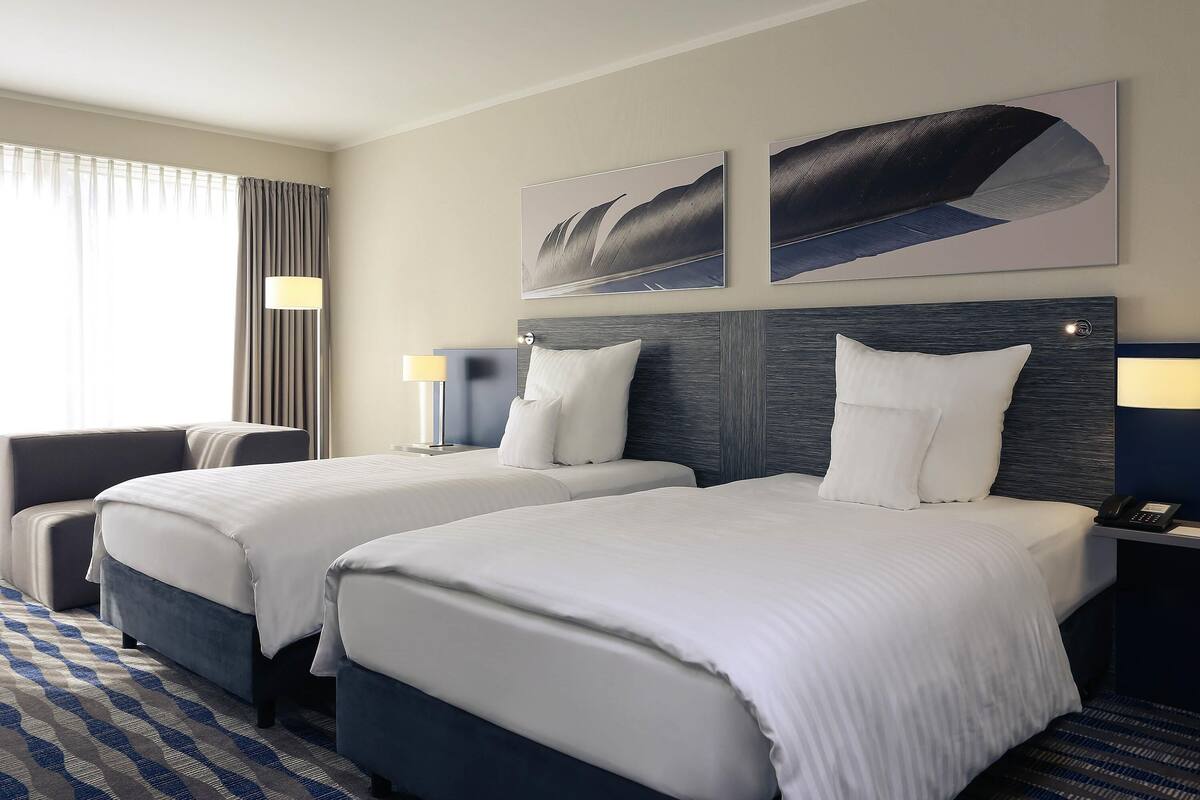 Een hotelkamer met twee bedden, een groot raam met gordijnen, een schilderij aan de muur en een kamerbreed tapijt met patroon.