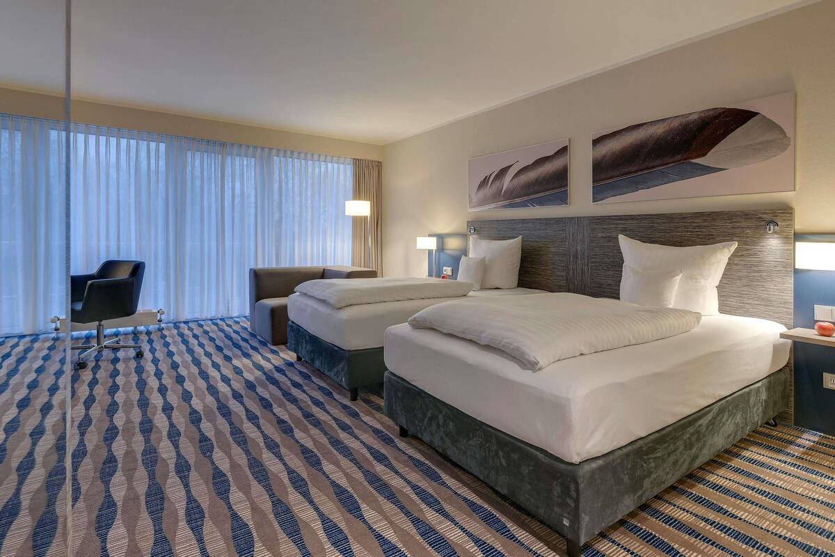 Quarto de hotel com uma cama grande, duas mesas de cabeceira, uma escrivaninha com cadeira e uma janela ampla com cortinas.