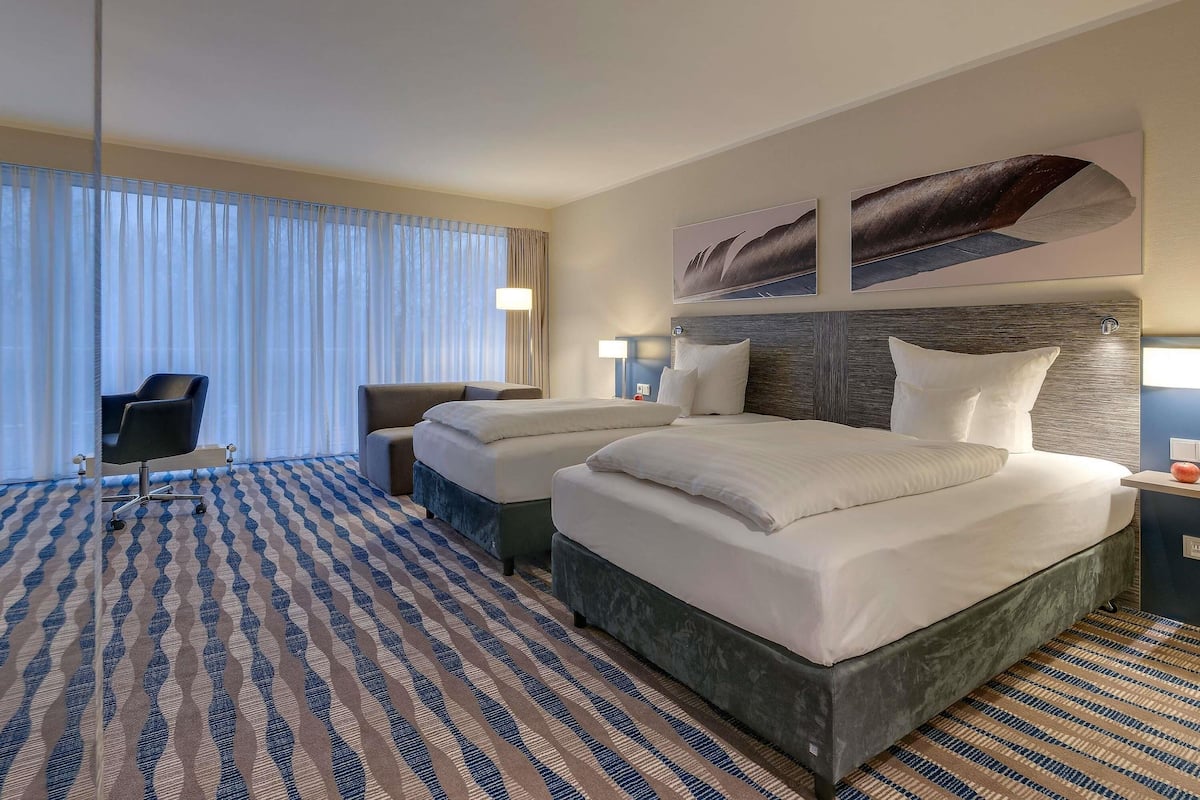 A hotel room with a large bed, two bedside tables, a desk with a chair, and a large window with curtains.