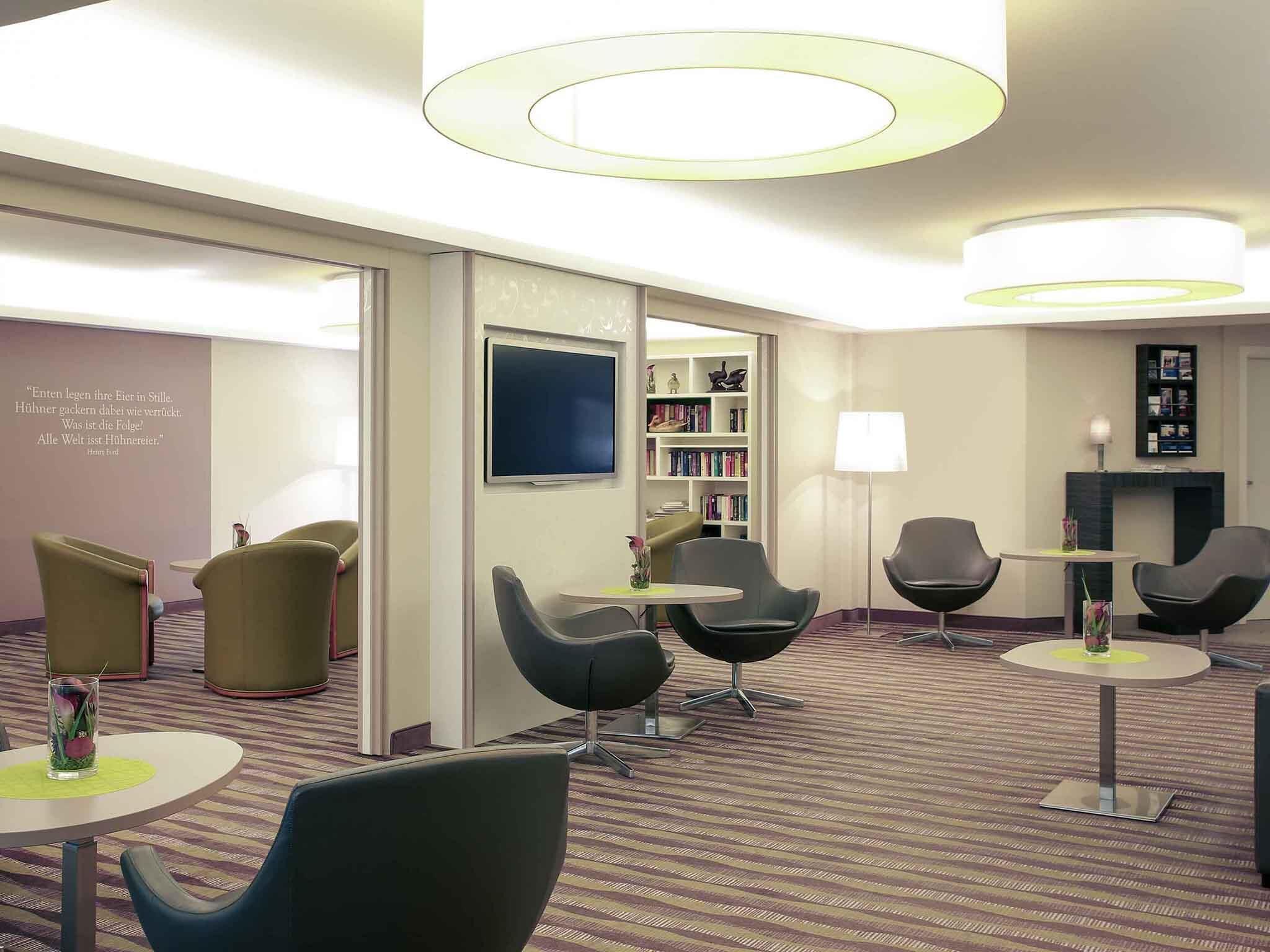 A modern hotel lobby with round tables, comfortable chairs, a bookshelf, a flat-screen TV, and a fireplace.